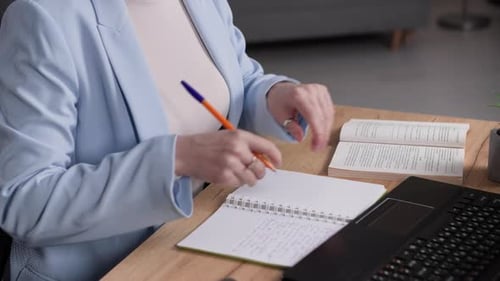 Young Woman Works in Office and Makes Notes in a Notebook and Works on Laptop While Sitting at Table
