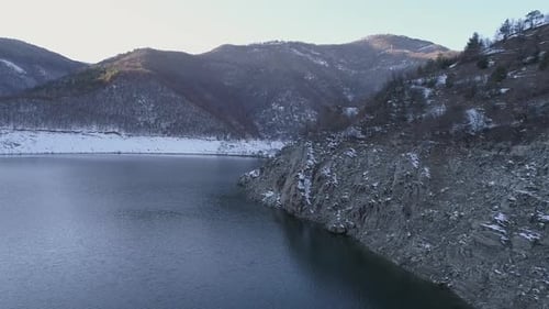 AERIAL: Flying above high rocky shore of a mountain lake on its shores there are hills covered with