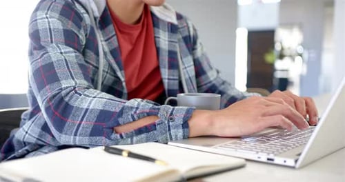 Tippt auf Laptop, Mann arbeitet von zu Hause aus mit Kaffee und Notizbuch in der Nähe