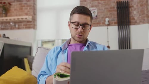Attractive Young Man Freelancer Sitting on Sofa Use Laptop Talk and Write in a Notebook Stay at Home