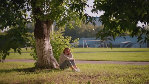 Thoughtful Woman Sitting Under Tree in Park with Stadium Background