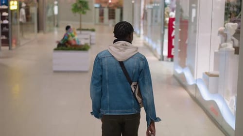 Young Black Man Walking Down Shopping Mall Corridor with Shoulder Bag