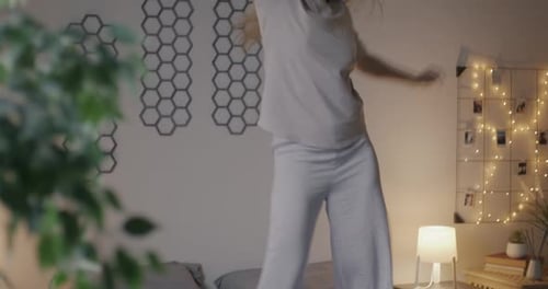 Happy Young Woman Dancing on Bed Listening to Music with Headphones Having Fun in Bedroom in Evening