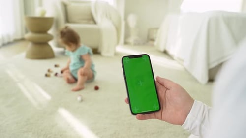 Junge Mutter kauft online auf der Website mit dem Greenscreen des Smartphones ein, während sie mit dem Kind spielt. Frau trägt