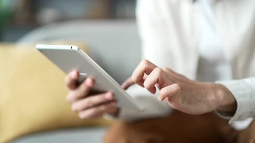 Close up of male hands holding digital tablet sitting on sofa in living room at home. Man browses