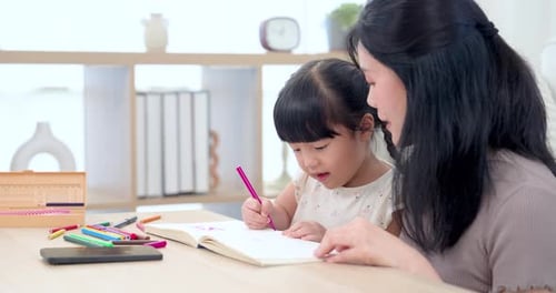 Asiatische alleinerziehende Mutter coacht Tochter im Vorschulalter beim Zeichnen in einem offenen Arbeitsbuch, Kind bewegt sich, rosa Stift