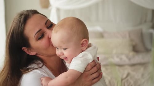 Mother Hugging and Kissing Smiling Infant in Bedroom