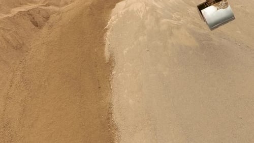 An aerial view of an excavator scoop digging into a sand pile, capturing the texture of the sand.