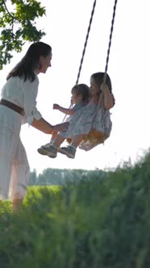Vertical of Mom Swings Her Two Happy Daughters on a Rope Swing in the Middle of a Green Field