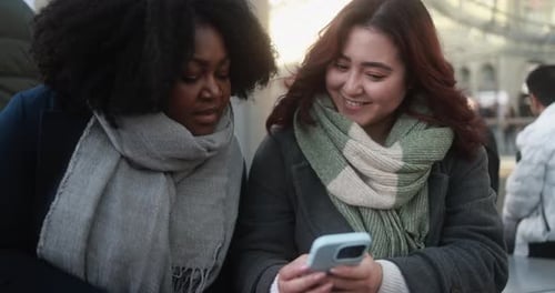Young multiracial women using mobile phone while waiting together at bus station