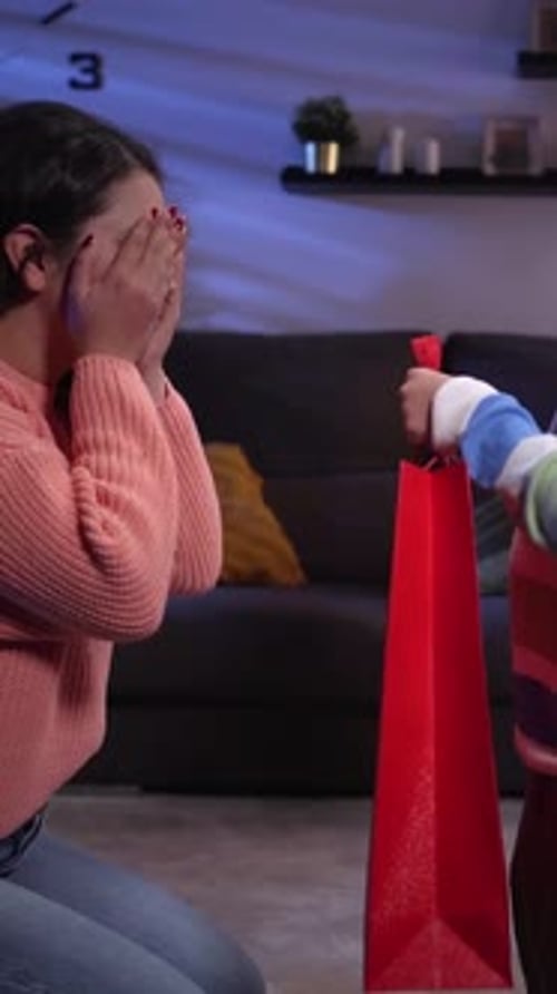 El hijo le da un regalo a su madre mientras ella lo espera con los ojos cerrados en la sala de estar