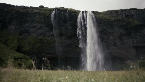 Wide dolly shot of an absolutely stunning scene of a waterfall, cliffs, and green field. Seljalandsf