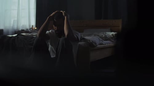 Man Sitting on Floor Near Bed in Dark Room