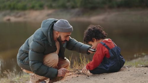 Un jeune père et son fils aux cheveux bouclés examinent la nature avec une loupe au bord du lac