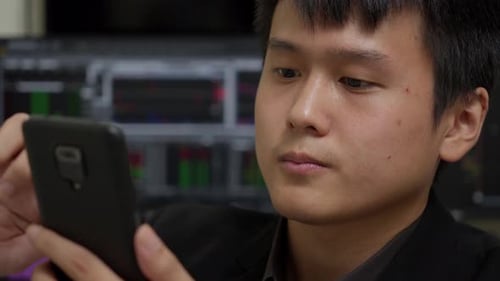 Young Asian man smiling while looking at smartphone, office environment with stock market charts in