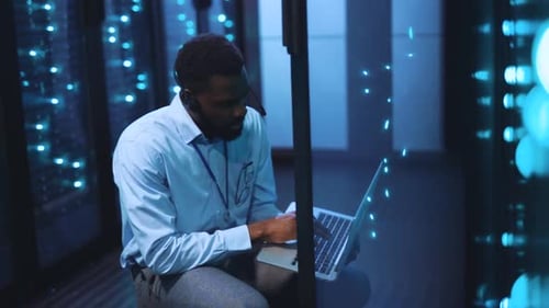 A Man Works Intently in a Server Room with His Laptop and Headphones