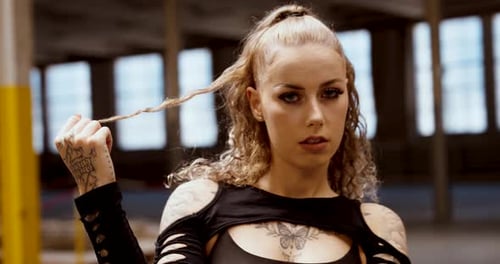 Tattooed woman playing with hair while looking into camera – confident close-up in industrial settin