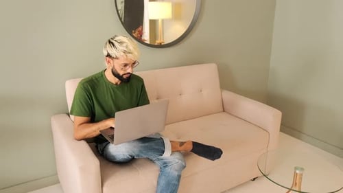 Young blond man with a beard works on a laptop while sitting in a cozy living room