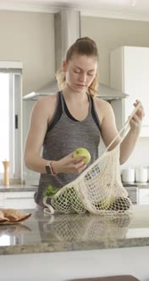 Vídeo vertical: Mujer levantando las asas de una bolsa de malla, sacando e inspeccionando una manzana en la cocina para comer
