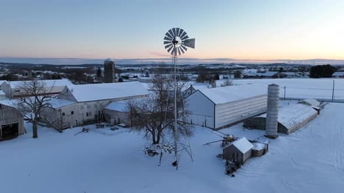 Dawn lights up a snow-covered farm with barns, a silo, and a classic windmill, embodying a serene ru