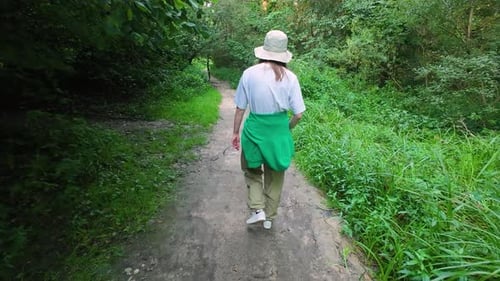 A young girl walks along a forest path among trees and grass in a summer forest.