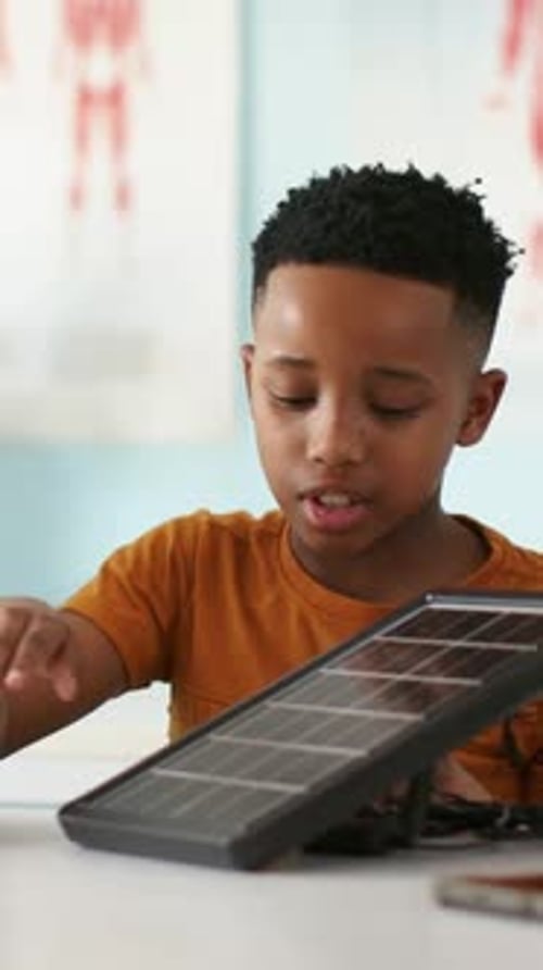 Vertical video: Leaning, youth in orange tee adjusting solar panel in classroom, showing panel use