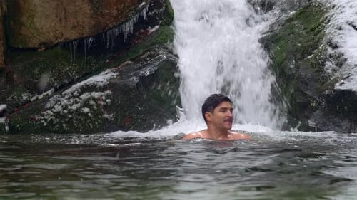 Handsome man ice swimming in subzero cold river by the beautiful waterfall in snowy mountains during