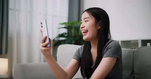 A young woman is sitting on a cozy sofa, happily video calling someone.
