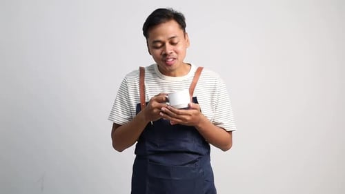 A male coffee shop waiter wearing an apron smells the aroma of coffee in a cup with a smile.