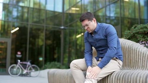 Businessman suffers from painful severe knee joint pain sitting on a bench on the street near office