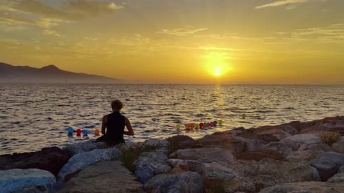 Lonely Woman Sitting On The Rocks On The Beach Watching The Sea At Summer Sunset