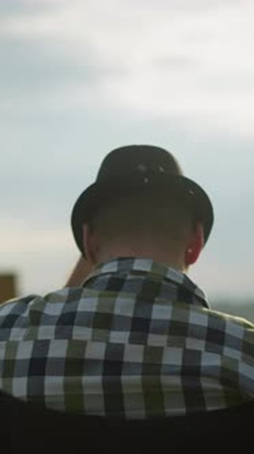 Man in Hat Adjusting It While Sitting in Field with Woman Painting in Background