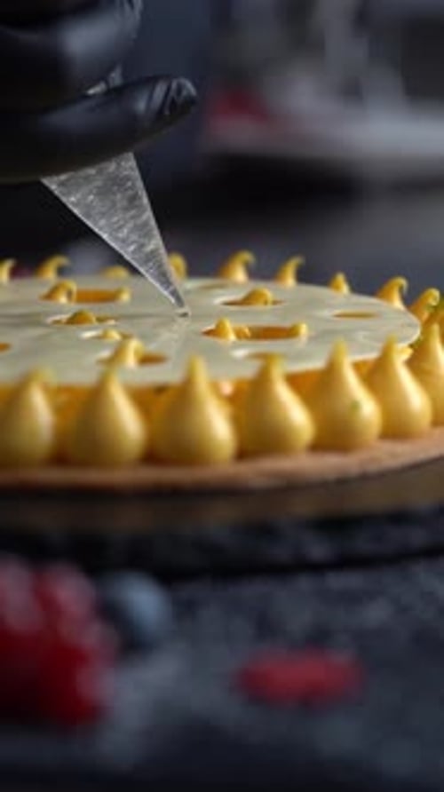 A chief puts smooth cream drops on a modern yellow cake dessert, close up shot