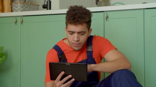Man with Tablet Sits on Floor Near Kitchen Cabinets