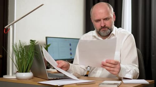 Man Reviews Documents at Desk, Frustrated