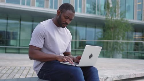 Un homme afro-américain indépendant utilisant un ordinateur portable en train de taper du texte discutant dans une rue urbaine d'une ville Guy ayant