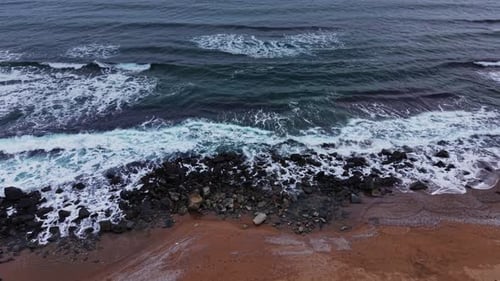 Waves crash on rocky shore near sandy beach during cloudy afternoon