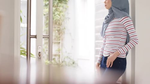 Portrait of thoughtful biracial woman in hijab at home by window with copy space, slow motion
