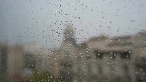 Vue rapprochée au ralenti de gouttes de pluie vues à travers une fenêtre lors de fortes pluies, avec une vue urbaine