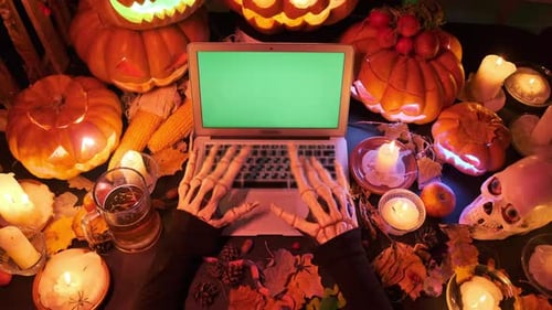 Laptop with green screen on typing bony hands promo party invitation next to pile pumpkins lanterns
