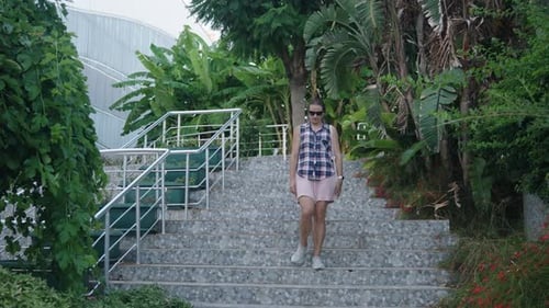Mulher desce uma grande escadaria de pedra, ladeada por folhagem tropical e um edifício moderno.
