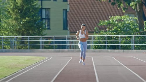 Happy and Smiling Fitness Woman in Light Blue Athletic Top and Leggings Jogging in a Stadium. She i