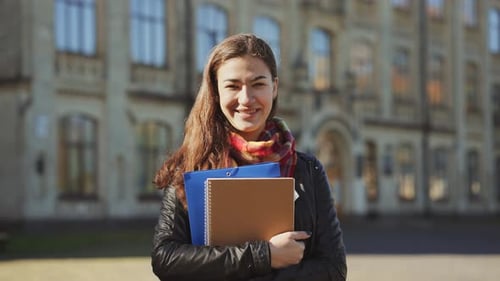 Smiling female student with books in hands standing in front of college building