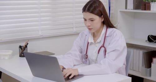 Asian female doctor wearing white coat and stethoscope working on laptop in clinic office, focused