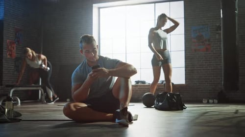 Handsome and Masculine Athletic Young Man is Using a Smartphone while Sitting on a Floor in a Loft