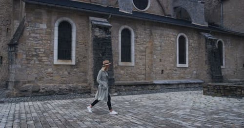 Stylish Young Woman Wearing Coat and Hat Walks Through Old European Church Yard. Beautiful Girl and