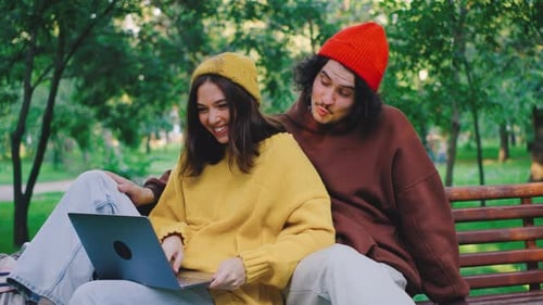 A Young Couple is Comfortably Seated on a Park Bench Deeply Engaged with Their Laptop While Enjoying