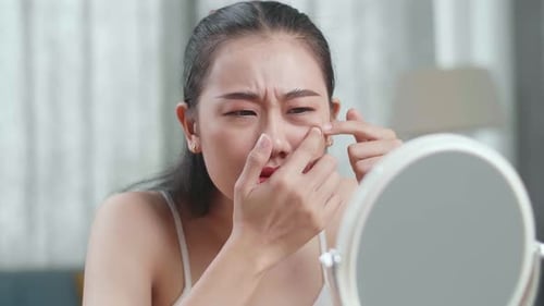 Close Up Of Worried Young Asian Woman Looks In The Mirror And Popping Pimples While Applying Makeup