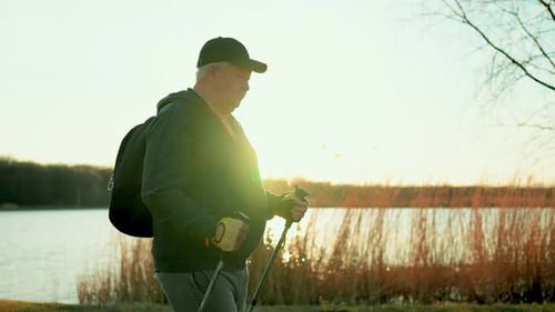 Active Handsome Elderly Man with Nordic Walking Sticks Walks Forward Through Along the Lake at