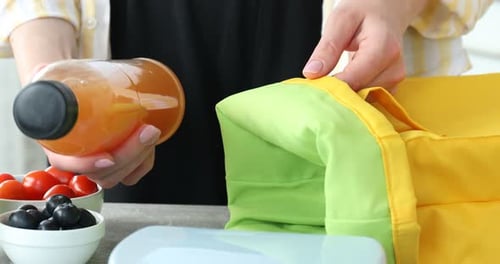 Mujer poniendo una botella de jugo y una lonchera en una bolsa en una mesa gris en el interior, primer plano